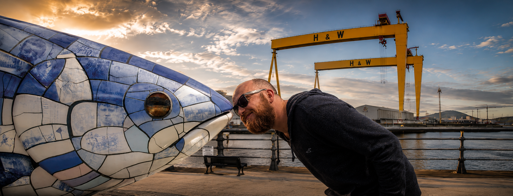 A man wearing sunglasses and a dark hoodie leans in closely to inspect the blue and white mosaic tile pattern of a large fish sculpture at a harbor, with yellow H&W cranes and a cloudy sky in the background.