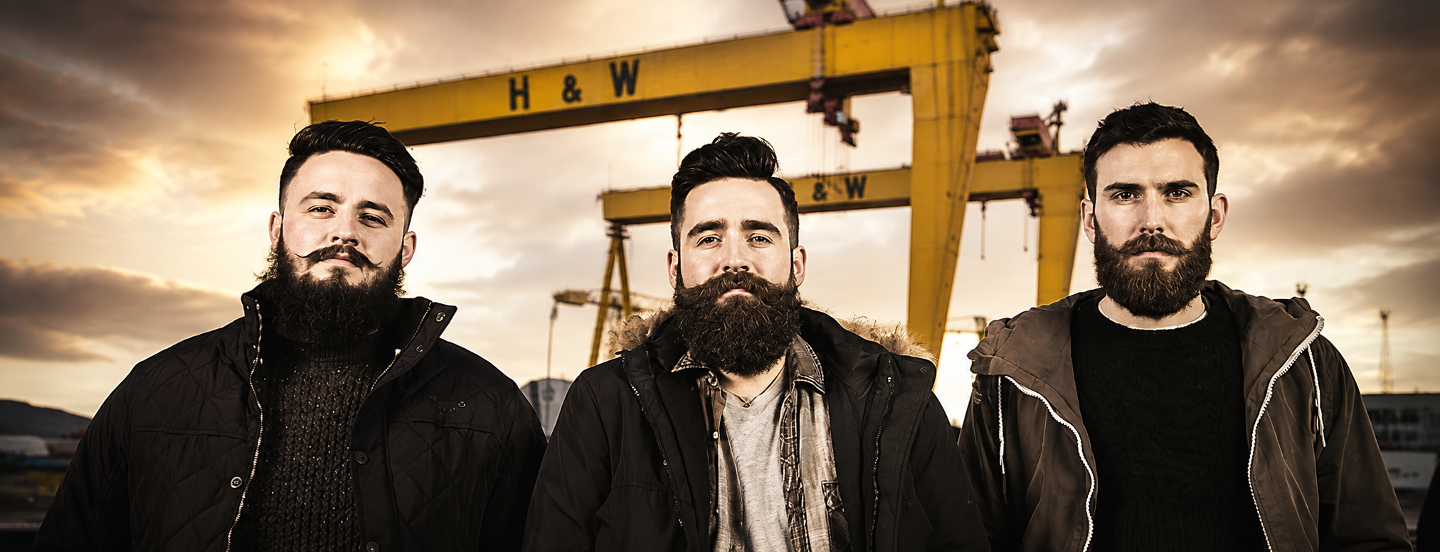 Three bearded men wearing dark jackets stand in front of a large yellow industrial crane against a dramatic sunset sky.