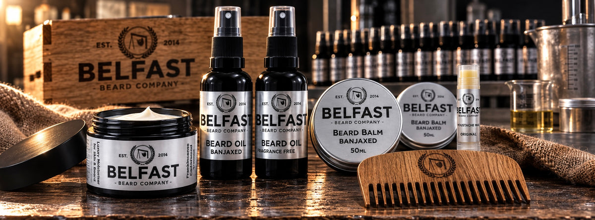 Belfast Beard Company beard care products arranged on a wooden surface, including beard oil bottles, beard balm jars, a lip balm tube, a wooden comb, and a branded wooden box in the background.